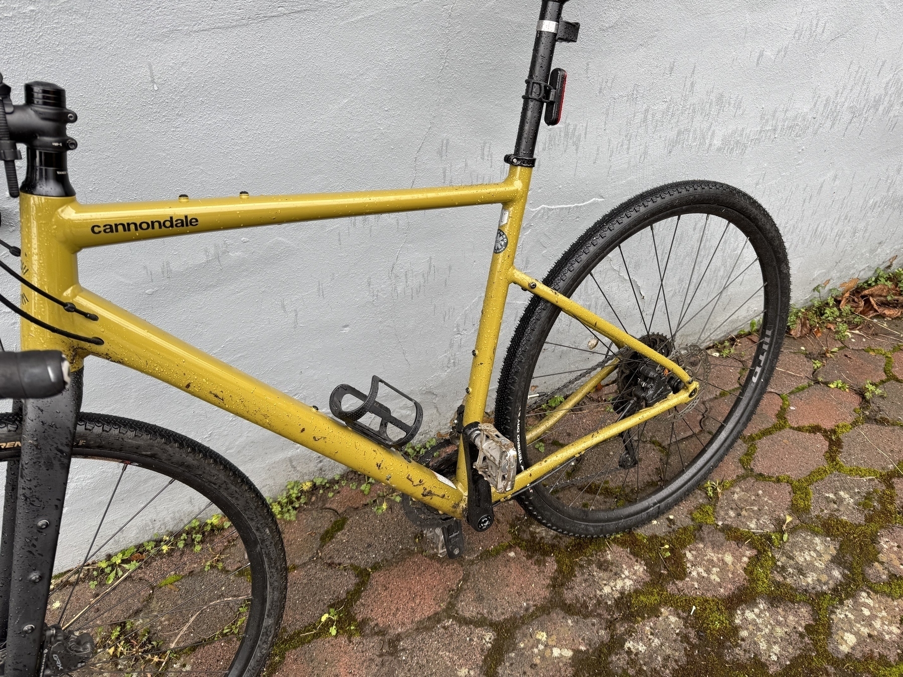 A wet and dirty Cannondale bicycle is parked against a light-colored wall on a brick surface.